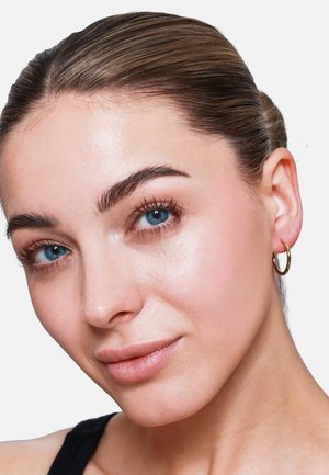 Boucle d'oreille en or dans l'oreille d'un modèle, réfléchissant la lumière. Texture de peau lisse, yeux bleus clairs et maquillage léger mettent en avant.