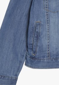 Džínová bunda v světle modré barvě, klasický design, viditelné švy a boční kapsa. Materiál má texturovaný povrch.