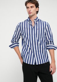 Μπλε και λευκή ριγέ πουκάμισο με γιακά και μία τσέπη στο στήθος, διαθέτοντας σηκωμένα μανίκια και λεπτή γραμμή.