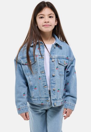 Jeune fille aux longs cheveux bruns portant une veste en jean bleu clair et un jean, avec un t-shirt blanc en dessous, debout devant un fond blanc.