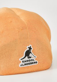 Căciulă tricotată portocalie, cu un model texturat, având un logo negru Kangol și "J.Lindberg" pe un fundal alb.