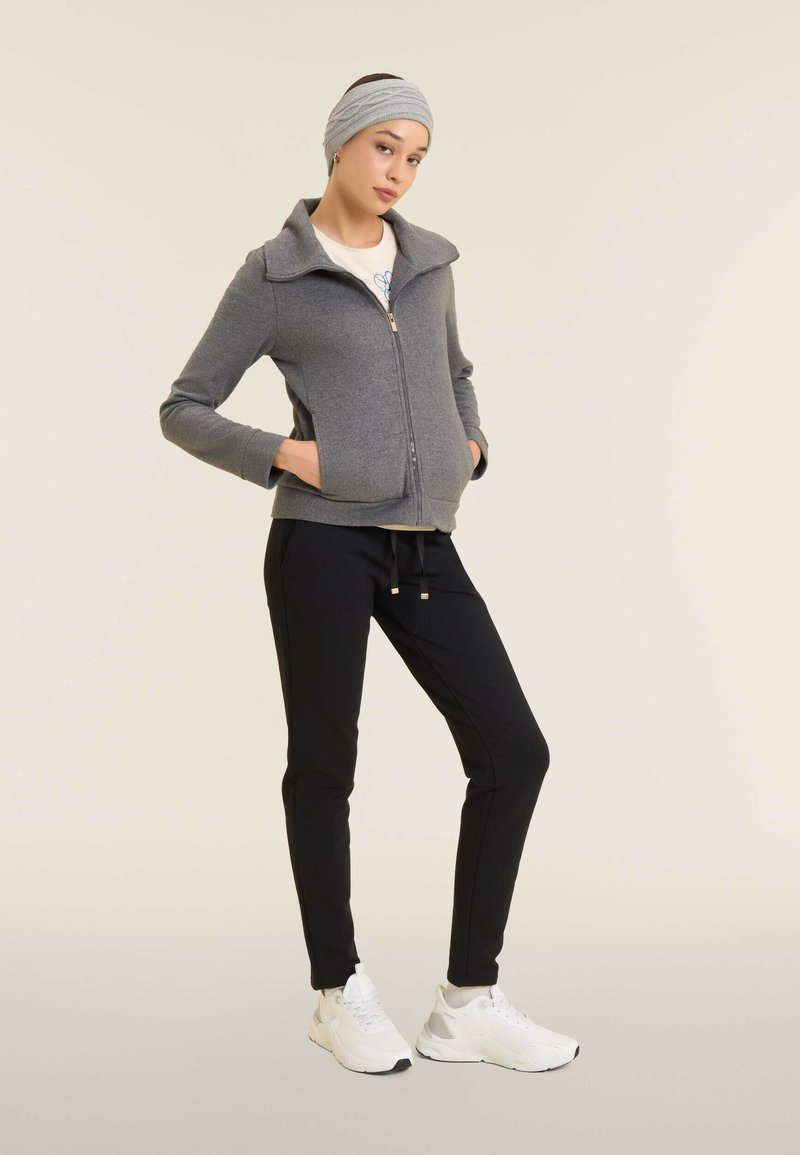 Giacca grigia con zip e colletto, pantaloni neri a gamba stretta e sneakers bianche. La fascia per capelli è grigia e la giacca presenta tasche laterali.