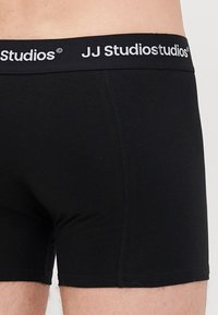 Schwarze Boxer-Shorts aus weichem, dehnbarem Material. Mit einem breiten elastischen Bund, auf dem das Logo "JJ Studios" in Weiß prangt. Sauberes Design ohne Muster.