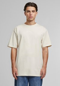 Beige bomulds-t-shirt med korte ærmer, rund halsudskæring og hvidt broderet logo på brystet. Blød tekstur med en afslappet pasform.
