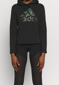 Hoodie noir à manches longues avec cordons réglables, featuring un logo Adidas en camouflage. Porté avec des leggings en maille texturée noirs.