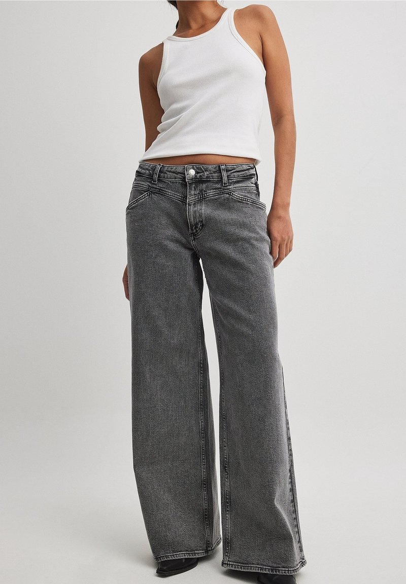 Personne portant un haut blanc sans manches et un jean noir ample taille haute, debout devant un fond uni.