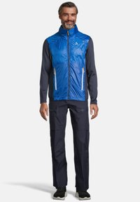 Homme debout, portant un gilet sans manches zippé bleu par-dessus une chemise à manches longues sombre, un pantalon sombre avec des poches zippées et des baskets noires.