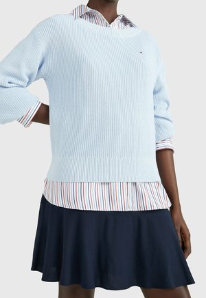 Personne portant un pull en tricot bleu clair sur une chemise rayée à boutons et une jupe bleu marine, debout devant un fond clair uni.