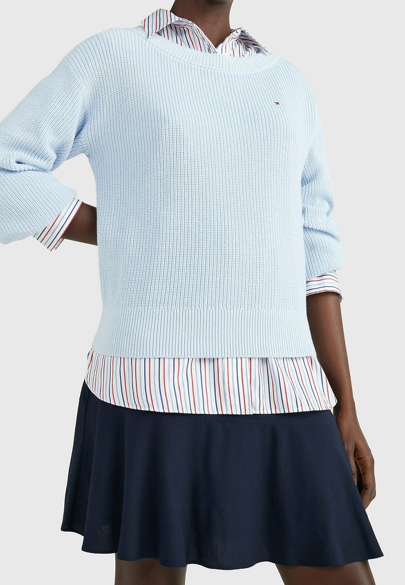 Personne portant un pull en tricot bleu clair sur une chemise rayée à boutons et une jupe bleu marine, debout devant un fond clair uni.