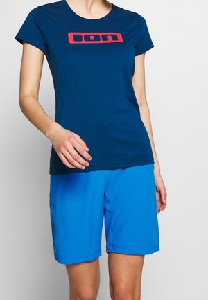 Person trägt ein eng anliegendes dunkelblaues T-Shirt mit rotem Logo und lockere, hellblaue Shorts, steht vor einem einfarbigen hellen Hintergrund.