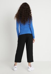 Una persona con lunghi capelli ricci si trova di spalle, indossando una maglietta a maniche lunghe blu, pantaloni neri cropped e sneakers bianche FILA.