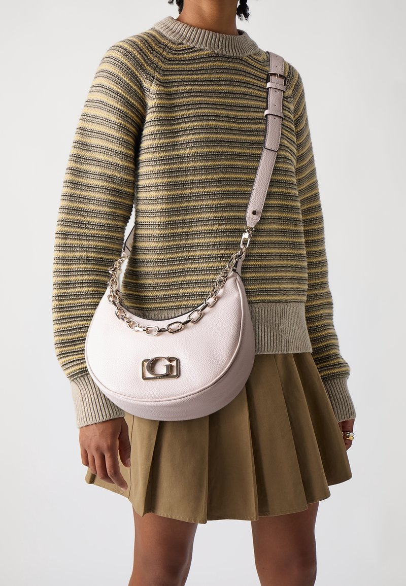 Personne portant un pull rayé jaune et gris et une jupe plissée kaki, portant un sac à main demi-lune rose clair avec chaîne et logo "G".