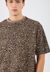 Läiskäkuvioinen lyhythihainen t-paita ruskeana ja beigeenä, jossa on pyöreä beige kaula-aukko ja pehmeä rakenne. Ainulaatuinen eläinkuvio ympäriinsä.