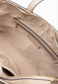 Beige tote-väska med ett inre fack och slät tygfoder. Guldzipperdetaljer och svarta kantdetaljer längs kanterna.