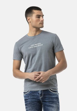 Junger Mann trägt ein graues T-Shirt mit dem Text "Unlimited Cipo and Baxx", kombiniert mit blauen Jeans, schaut zur Seite.