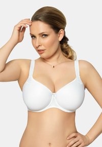 Donna con capelli castano chiaro raccolti in una coda di cavallo bassa, che indossa un reggiseno bianco con ferretto, toccandosi i capelli con una mano e appoggiando l'altra sui fianchi.