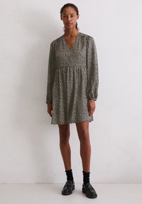 Sukienka w kwiatowy wzór w ciemnozielonym i kremowym kolorze, z dekoltem w serek, długimi rękawami i marszczonym pasem, zestawiona z czarnymi butami i skarpetkami.