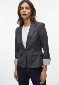 Gray tailored blazer with a single button, two front pockets, and striped cuffs. Material is smooth with structured fit. Worn over a white shirt.