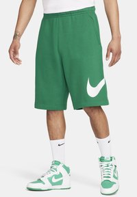 Muž oblečený v zelených šortkách Nike s bílým logem swoosh, v kombinaci s bílými ponožkami Nike a zeleno-bílými teniskami Nike, stojící na bílém pozadí.