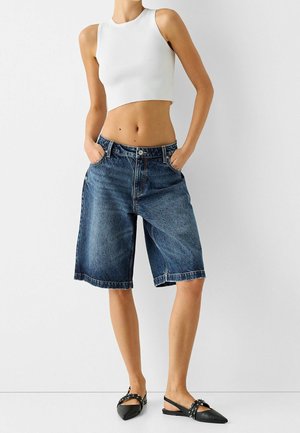 Shorts en denim bleu foncé avec une coupe décontractée, cinq poches et un ourlet retroussé ; associés à un top court en rib blanc et des chaussures noires.