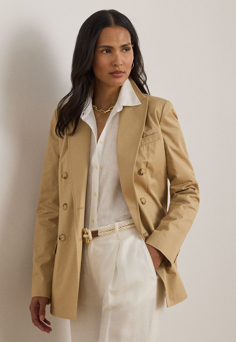 Γυναίκα με μακριά σκούρα μαλλιά φορά μπεζ σακάκι διπλής γραμμής, λευκό πουκάμισο με κουμπιά, λευκό παντελόνι και χρυσή αλυσίδα, στέκεται πλευρικά.