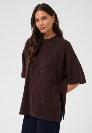 Femme aux longs cheveux foncés portant un pull ample à manches courtes marron foncé et un pantalon foncé, debout devant un fond clair uni.