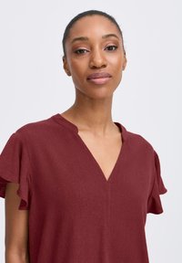 Blusa bordeaux con scollatura a V e maniche arricciate, realizzata in un tessuto texture. Il taglio è rilassato, con decorazioni minime.