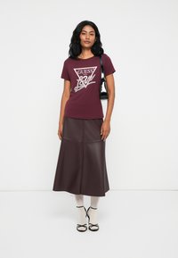 Vrouw in een bordeauxrode Guess graphic t-shirt, een donkerbruine leren midi-rok, witte kousen, zwarte sandalen en een zwarte schoudertas.