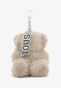 Llavero de oso de peluche beige y esponjoso con una cadena y broche de metal plateado, que incluye una etiqueta colgante de plata con la palabra "TOUS" en letras negras.