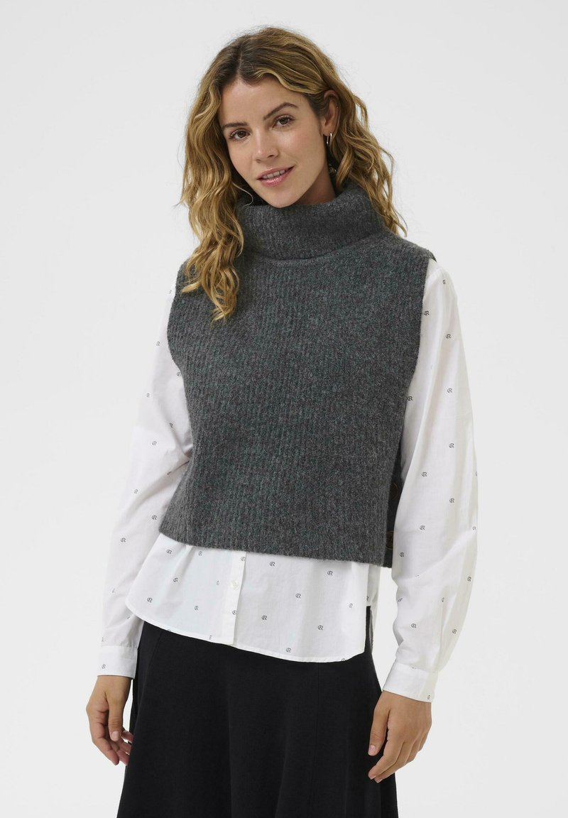 Sivi rebrasti turtleneck prsluk izrađen od mekog, teksturiranog materijala, nošen preko bijele košulje s malim uzorkovanim detaljima, u kombinaciji s crnom suknjom.