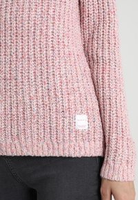 Różowy sweter z dzianiny o prążkowanej teksturze, zdobiony wielokolorowymi flekami. Zawiera małą białą metkę marki na dolnym lewym brzegu.