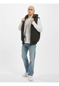 Gevoerde zwarte vest met een capuchon, ritssluiting, gedragen over een wit shirt met lange mouwen. Lichtblauwe jeans en witte sneakers maken de outfit compleet.
