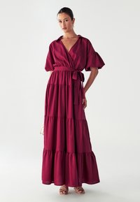 Dlouhé, patrové maxi šaty v tmavě burgundské barvě. Mají krátké volánkové rukávy a design s překrytím a zavinovacím páskem v pase. Hladká struktura látky.