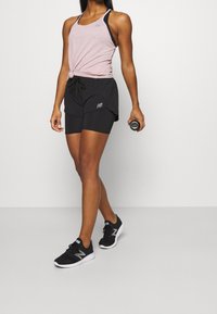 Kvinna står och håller en vattenflaska, iförd en rosa ärmlös topp, svarta lager shorts och svarta sportskor mot en vit bakgrund.