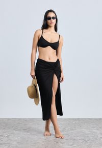 Top de biquíni preto com alças finas, combinado com uma saia preta tipo envelope com uma fenda frontal. Modelo segura um chapéu de palha e está descalça.