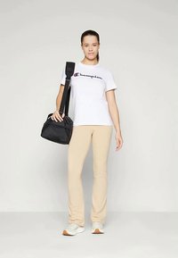 Jauna sieviete, ģērbusies baltā Champion t-kreklā, bēšos izmēros, baltās sporta kurpēs un ar melnu plecu somu, stāv uz vienkārša fona.