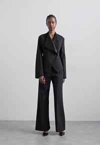 Sort skræddersyet jakkesæt med dobbeltradet jakke, brede revers og lange ærmer, parret med brede bukser og spidse hæle.