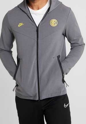Homme portant un sweat à capuche gris zippé avec des logos jaunes Nike et Inter Milan, mains dans les poches, pantalon Nike noir.