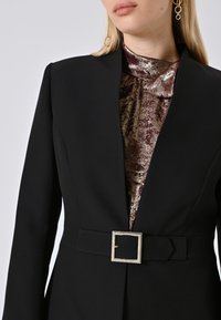 Donna che indossa un blazer nero con cintura sopra una maglia metallica viola lucida, con lunghi capelli biondi e orecchini a cerchio dorati.