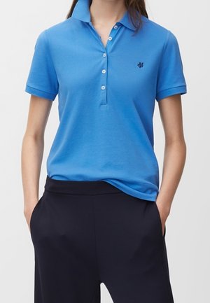Vrouw draagt een blauw kortärmelig poloshirt met knopen en zwarte broek, handen in de zakken, tegen een effen achtergrond.