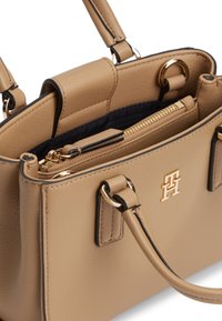 Μπεζ δερμάτινη τσάντα χειρός με διπλές λαβές, χρυσό φερμουάρ σε θήκη εσωτερικά και χρυστό μεταλλικό λογότυπο μπροστά, ορατή εσωτερική επένδυση σε σκούρο ύφασμα.