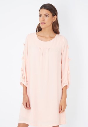 Vestido de color rosa claro, hasta la rodilla, con escote redondo, pliegues en la parte delantera y mangas de tres cuartos con acentos de pompones. Tela suave.