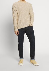 Osoba oblečená v béžovém texturovaném svetr s dlouhým rukávem, tmavých úzkých džínech s ohrnutými nohavicemi a béžových vysokých teniskách stojící na bílém pozadí.