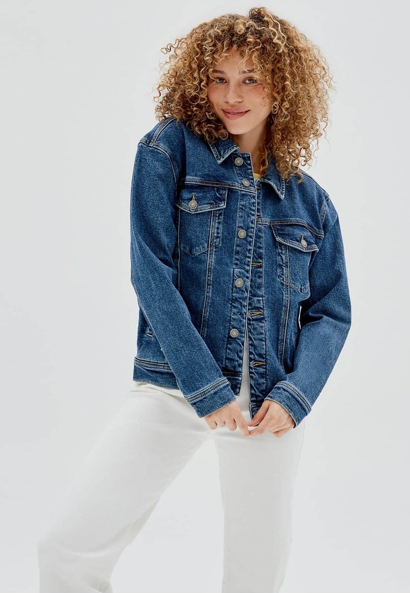 Giubbotto in denim di colore blu scuro con un taglio classico, apertura con bottoni, tasche sul petto e cuciture a contrasto. Presenta un colletto e maniche lunghe.