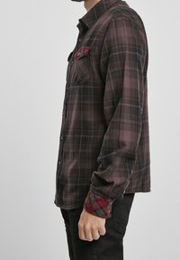 Chemise en flanelle à carreaux bruns avec fermeture à boutons, équipée d'une poche sur la poitrine et de poignets à carreaux rouges contrastants. Texture douce, coupe décontractée.