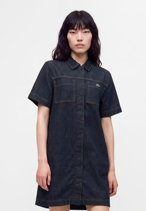 Giovane donna con lunghi capelli scuri indossa un vestito corto in denim scuro con maniche corte, tasche frontali e colletto, in piedi davanti a uno sfondo chiaro e uniforme.