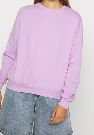 Personne portant un sweat-shirt ample lavande avec des poignets et un ourlet côtelés, associé à un short en denim bleu clair.