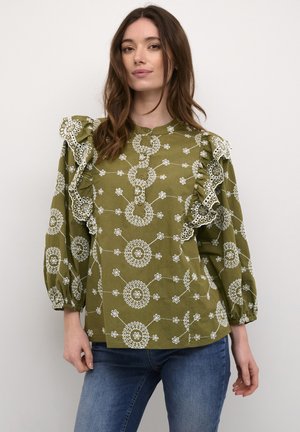 Vrouw in een olijfgroene blouse met witte geborduurde ronde patronen en gerimpelde schouders, in combinatie met blauwe jeans, staand tegen een witte muur.