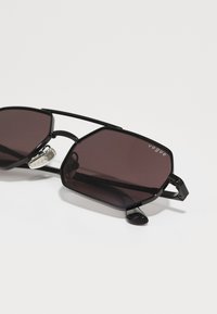 Óculos de sol hexagonais pretos com lentes escuras, estrutura de metal, almofadas nasais integradas e logótipo "Vogue" no canto superior esquerdo da lente.