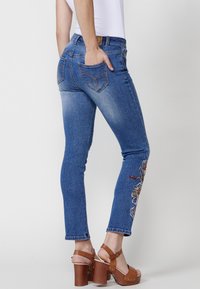 Blå denimjeans med smal passform, med blommönsterbroderi på vänster ben och två bakfickor. Bärs med bruna plattformssandaler.